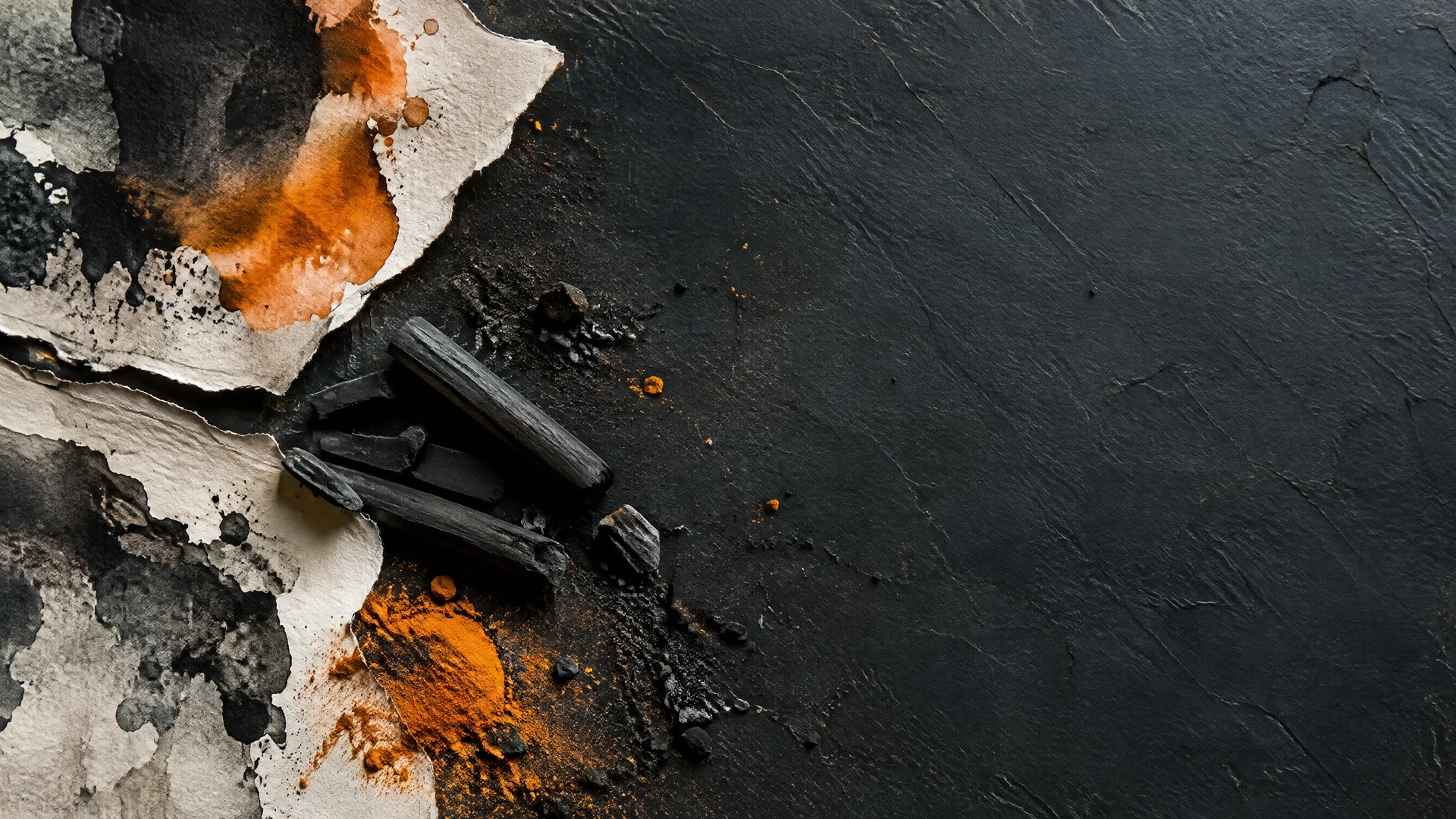 Torn paper edges with black ink and orange pigment, charcoal sticks and dust on a dark slate surface